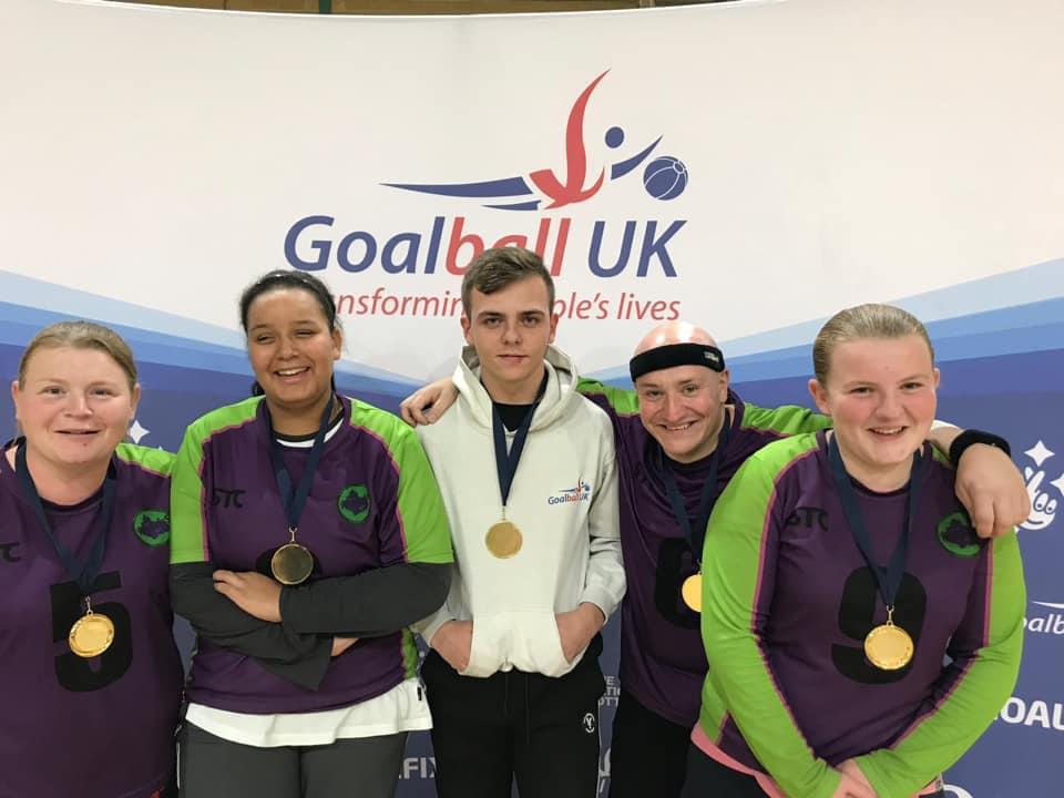Image shows Alex stood with members of the Croysutt Warriors team in front of a Goalball UK banner. She has a big smile on her face!