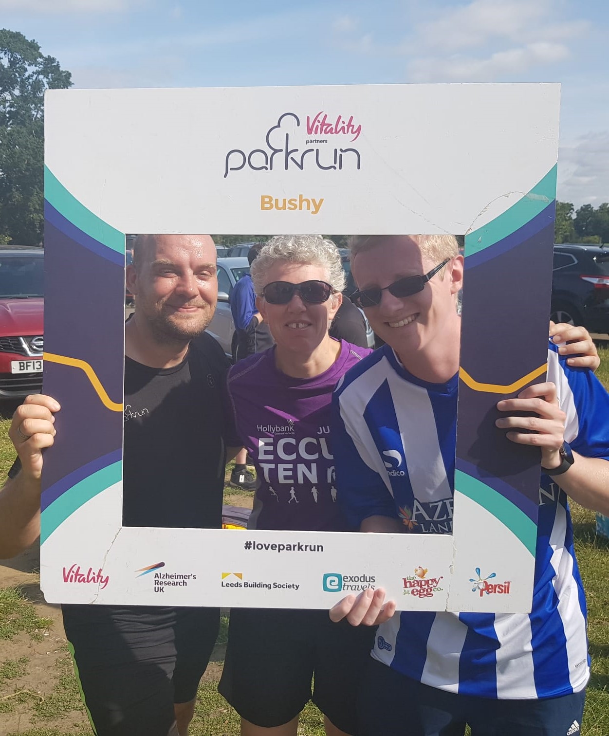 Phil, Kathryn and Stephen at Busy parkrun holding the selfie frame with the parkrun logo and Bushy on it