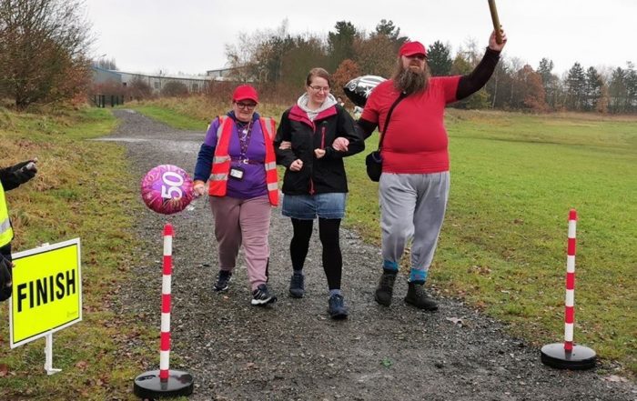 Chris Payne, holding aloft a 'golden baton' as he crosses the finish line at Poolsbrook parkrun together with Teresa (and a volunteer)