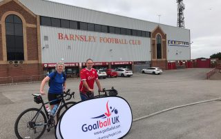 Kathryn and Phil, stood with their bikes, in front of a Goalball UK banner outside Oakwell Stadium