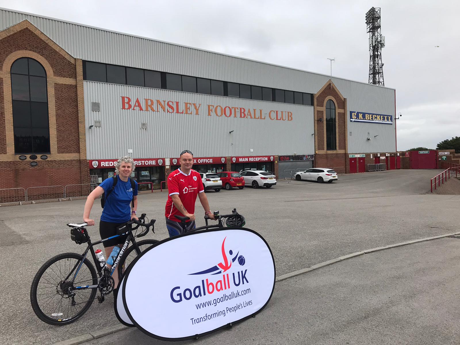 Kathryn and Phil, stood with their bikes, in front of a Goalball UK banner outside Oakwell Stadium