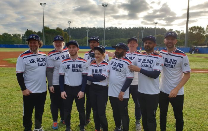 Team photo of the UK blind baseball team stood outside on a floodlit baseball diamond