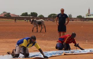 Chris Davies coaching goalball in a dusty field in Ghana. Meanwhile a cow is walking behind the court!