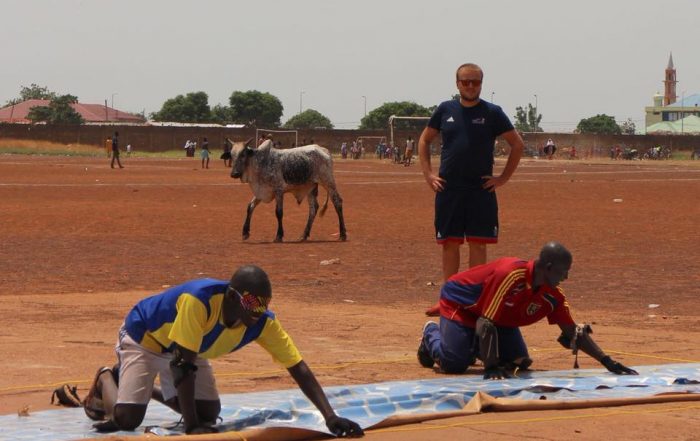 Chris Davies coaching goalball in a dusty field in Ghana. Meanwhile a cow is walking behind the court!