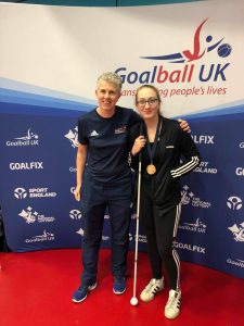 Samantha Gough and Kathryn Fielding stood together at a tournament in Sheffield in front of a Goalball UK banner.