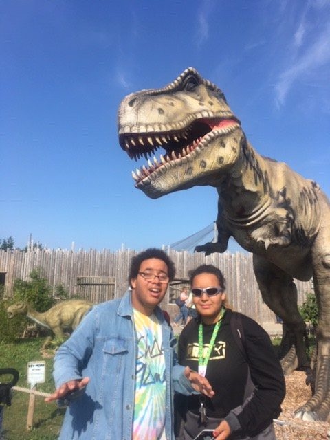 Image shows Alex stood with a friend in front of a T-Rex smiling at the camera!