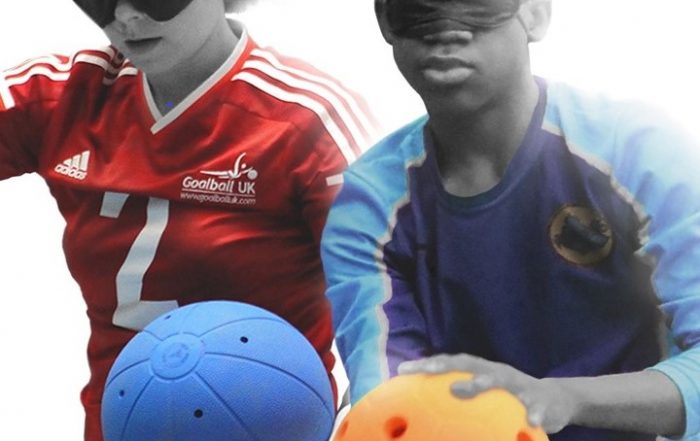 A Great Britain women's player holding a goalball, stood next to a young male player preparing to shoot an orange jingle ball.