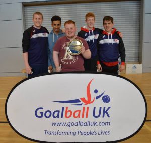 Image shows Ben stood holding a trophy along with members of the RNC goalball team. They are also stood with RNC and GB Womens coach Aaron Ford.