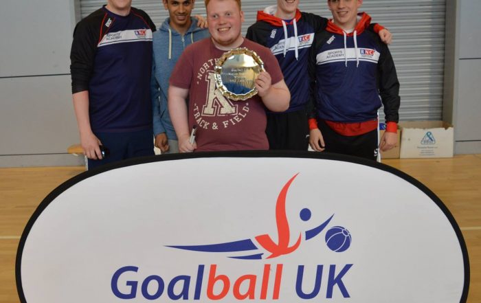 Image shows Ben stood holding a trophy along with members of the RNC goalball team. They are also stood with RNC and GB Womens coach Aaron Ford.