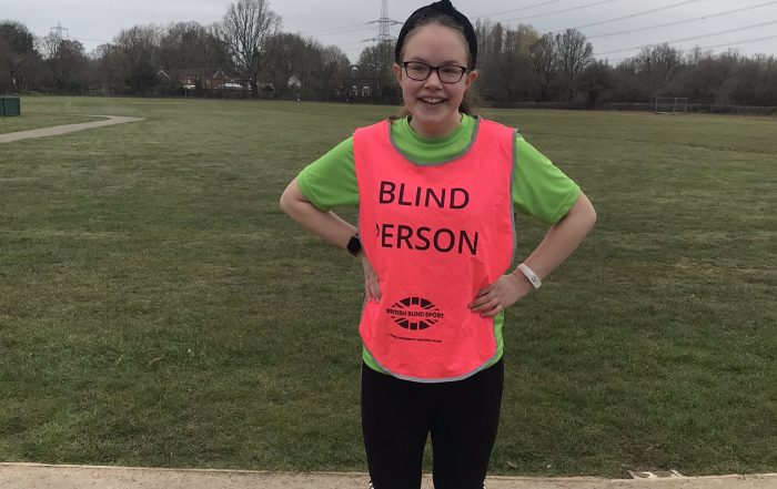 Keira stood smiling, wearing a 'Blind Person' bib, on a path by a grass field