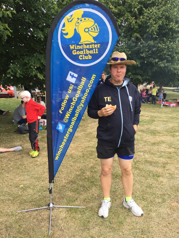 Image shows Tom Evison stood next to a Winchester Goalball Club banner with a burger at the 2017 Winchester BBQ