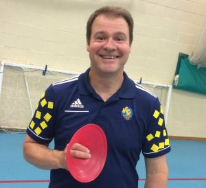 Image shows Tom Evison wearing his Winchester Goalball Club top, stood smiling at the camera and holding a red training cone