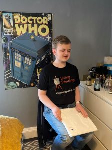 Louis, wearing a Living Paintings t-shirt, sat reading a 'Touch to See' book with a big Doctor Who poster on the wall behind him.