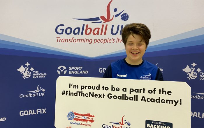 Freya Gavin stood holding a sign which reads "I'm proud to be a part of the #FindTheNext Goalball Academy" with a big smile whilst laughing!