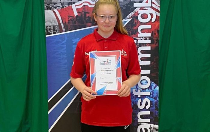 Ruby Hudson stood in front of a Goalball UK banner holding her referees course completion certificate after the 2021 Summer Camp.