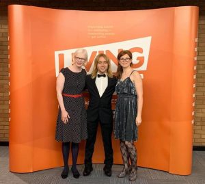 Sarah Leiter, Warren Wilson and Emily Watton stood together in formal attire in front of an orange background at an awards ceremony.