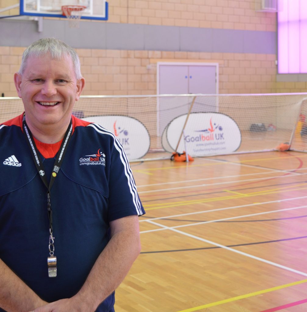 Robert Avery is stood on the edge of a goalball court smiling for the camera