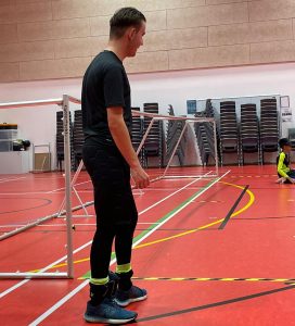 Jacob Hare stood next to the right corner of a goalball goal in a sports hall in a training session.