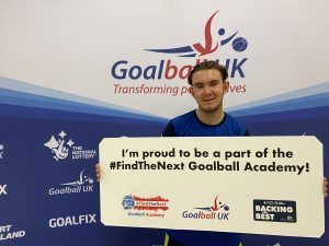 Jacob Hare stood in front of a Goalball UK banner with a sign saying "I'm proud to be a part of the #FindTheNext Goalball Academy" with a nice smile.