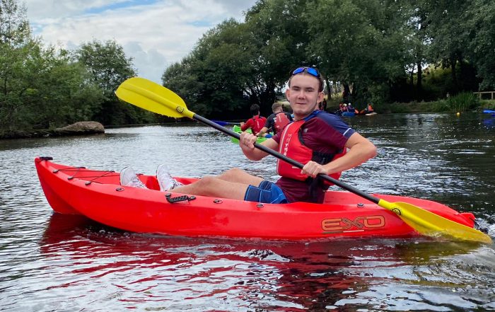 Jacob Hare in a red canoe with yellow canoe paddle on the water with large trees in the background.