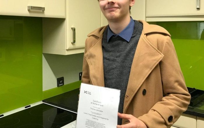 Tom Lancaster stood in a kitchen holding a certificate whilst wearing a beige coat.