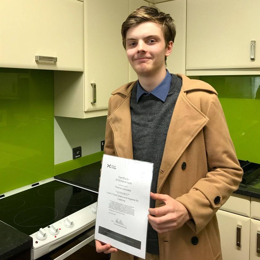 Tom Lancaster stood in a kitchen holding a certificate whilst wearing a beige coat.