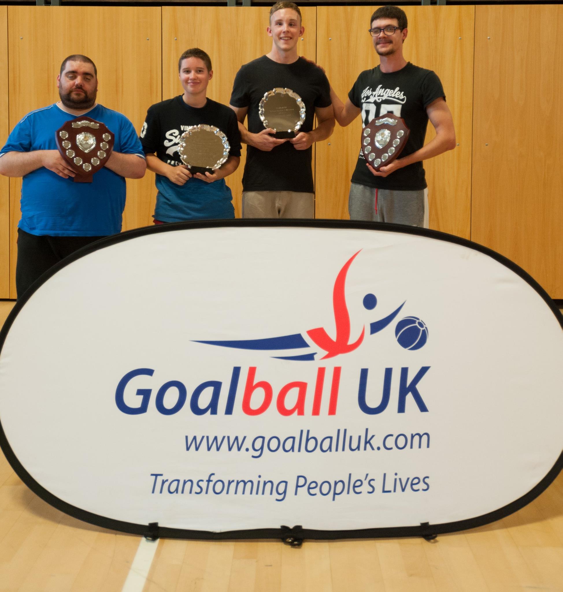 Matt Woof stood in a Lancashire Lions team photo in front of a Goalball UK banner. All 4 players are holding trophies!!