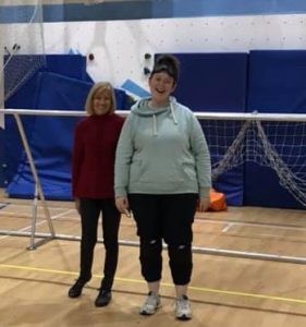 Two people standing on a goalball court in front of a goalball goal. 
