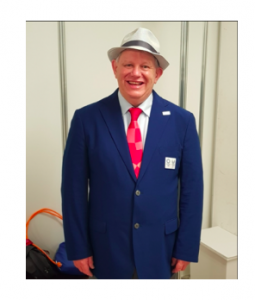 Robert Avery standing in his navy blue Tokyo 2020 suit with a red tie and a big smile!