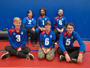 South Yorkshire Goalball team, with a row of three seated on the floor and three seated on a bench in their blue jerseys smiling.