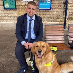 Jake Coles sat on a bench in a smart navy blue suit with his companion guide dog Jeffers sat next to him.