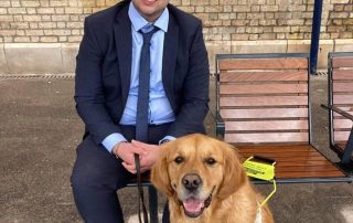 Jake Coles sat on a bench in a smart navy blue suit with his companion guide dog Jeffers sat next to him.