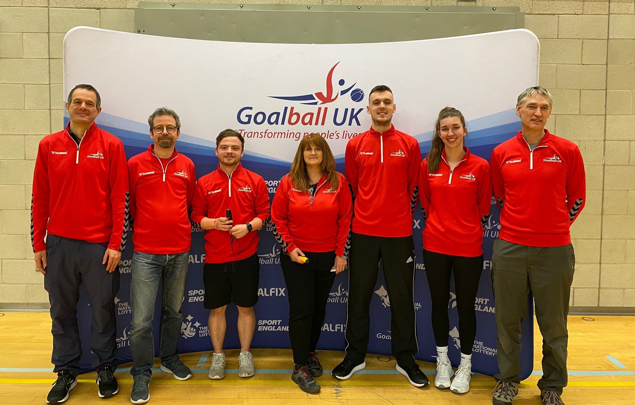 The 7 new Referees in their Goalball UK red fleeces in front of a Goalball UK banner. Left to right: Matthew Exley, Craig Meulen, Aaron Mitchell, Louise Gough, Stephen Reilly, Anna Martin, and Stuart Tapp.