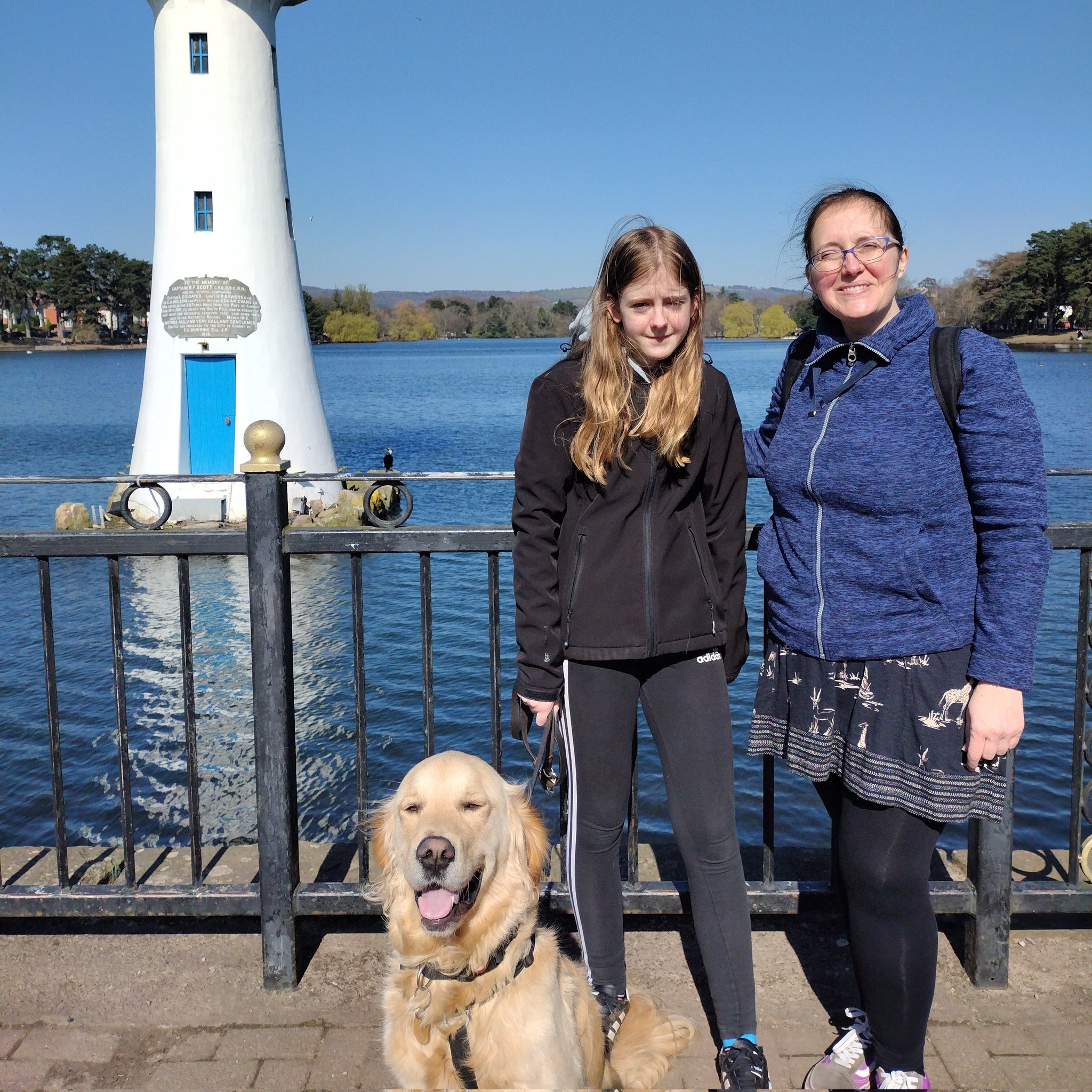 Catrin and Gwennan Young stood on a sunny pier with a lighthouse in the background. Catrin has got their furry companion buddy sat nearby.