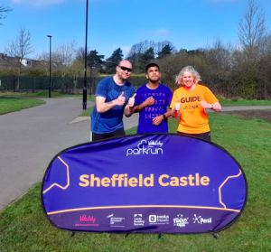 Phil, Yusuf, and Kathryn standing together in front of the parkrun Sheffield Castle banner all with thumbs up after completing their 5 k!