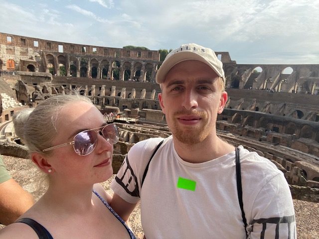Adam and his wife stood together at a colloseium, probably Rome! Adam is wearing a plain white t-shirt and a white cap.