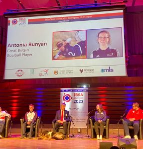 IBSA World Games launch with speakers sat on a stage. Goalball player Antonia Bunyan is on the stage with a profile of her on a large projector screen behind her.