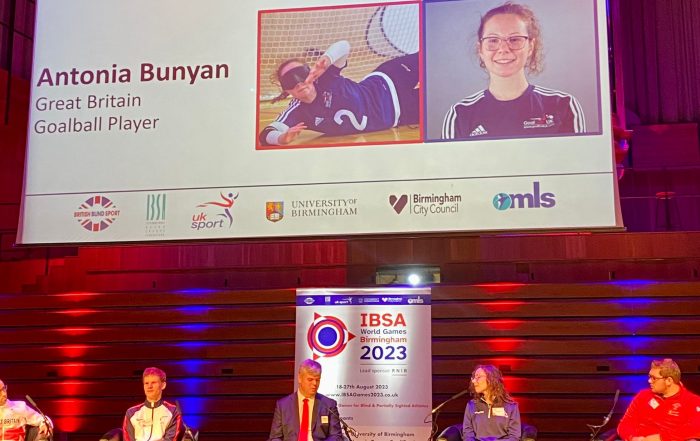 IBSA World Games launch with speakers sat on a stage. Goalball player Antonia Bunyan is on the stage with a profile of her on a large projector screen behind her.