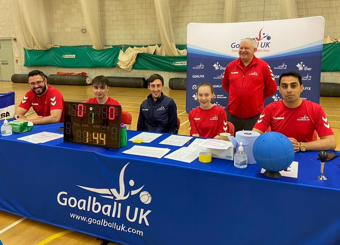 A picture of 4 Goalball UK activators in red shirts sat at the officials table with a referee stood behind them.
