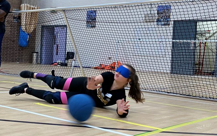 A girl at a past junior day on court diving to save the oncoming goalball whilst wearing soft blue eyeshades and a black top and black tracksuit bottoms.