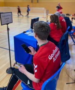 Harry Bainbridge wearing a bright red activator top whilst being a 10 second timer during a goalball game.