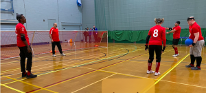 London Elephants warming up in a sportshall, all wearing their bright red jerseys. Russ, the coach is standing on the goaline to retrive the oncoming shots!