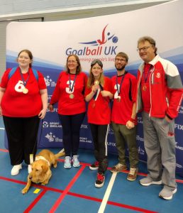 South Wales novice team group photo with 5 people stood together in bright red South Wales shirts with their bronze medals. A guide dog is laid down next to a player, pooped out after a busy day!