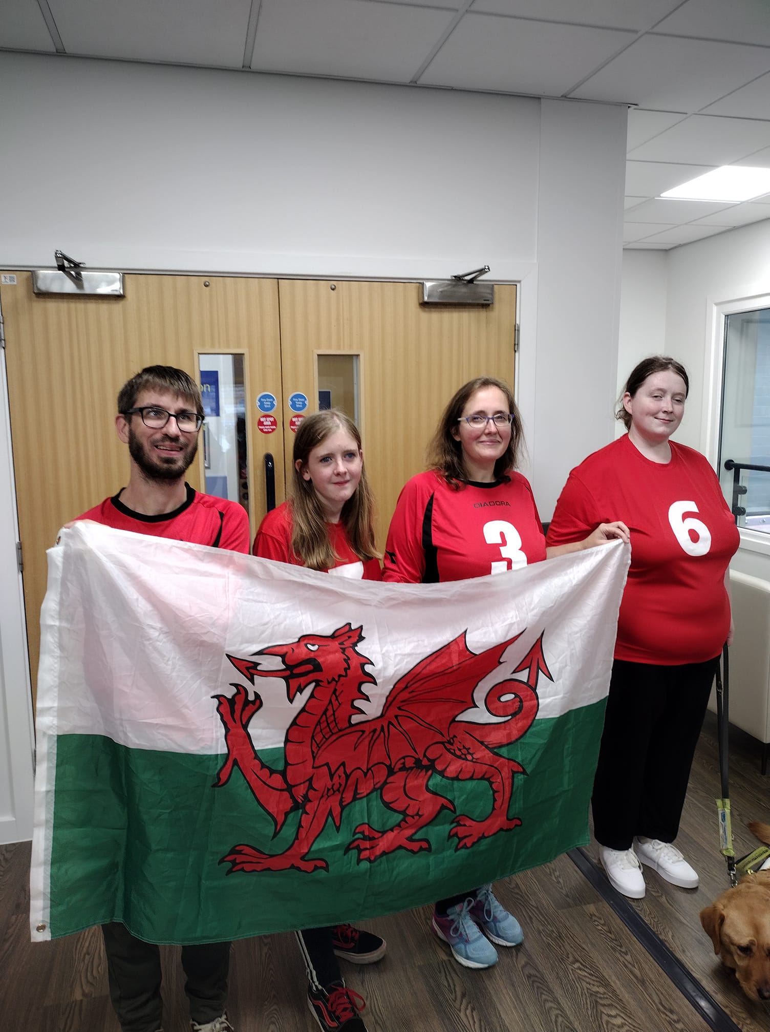 South Wales team of 4 players standing together holding a Welsh flag, with the top half white, bottom half green, and a red dragon in the middle.