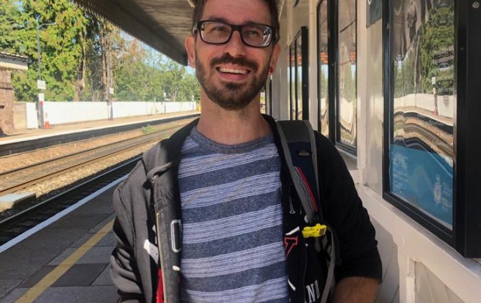 Marcus stood on a train station platform holding a costa coffee cup. Marcus is wearing glasses and a stripey blue top, with a thin grey jacket.