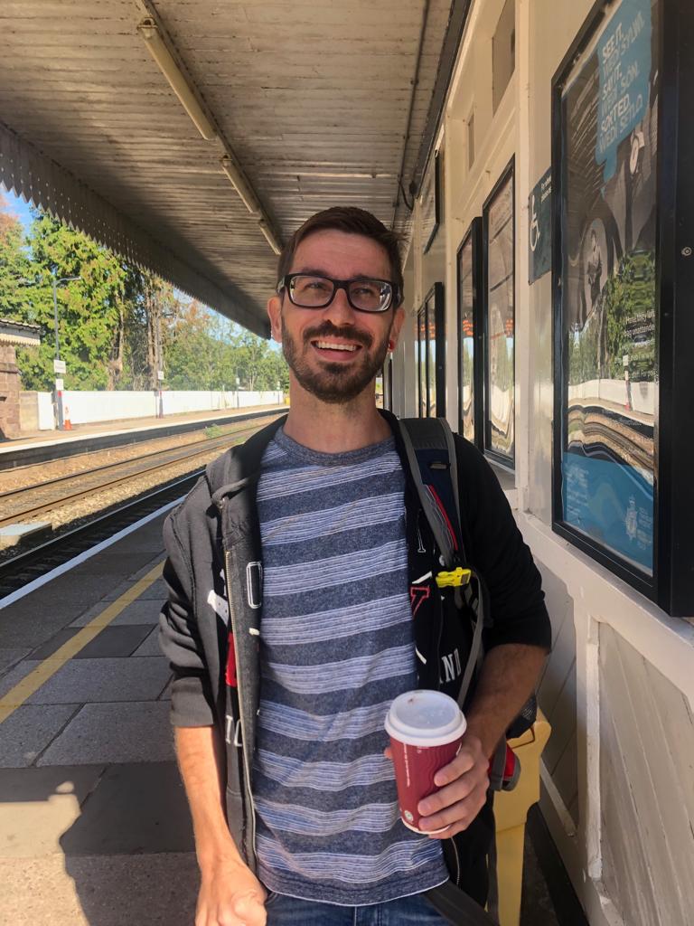 Marcus stood on a train station platform holding a costa coffee cup. Marcus is wearing glasses and a stripey blue top, with a thin grey jacket.