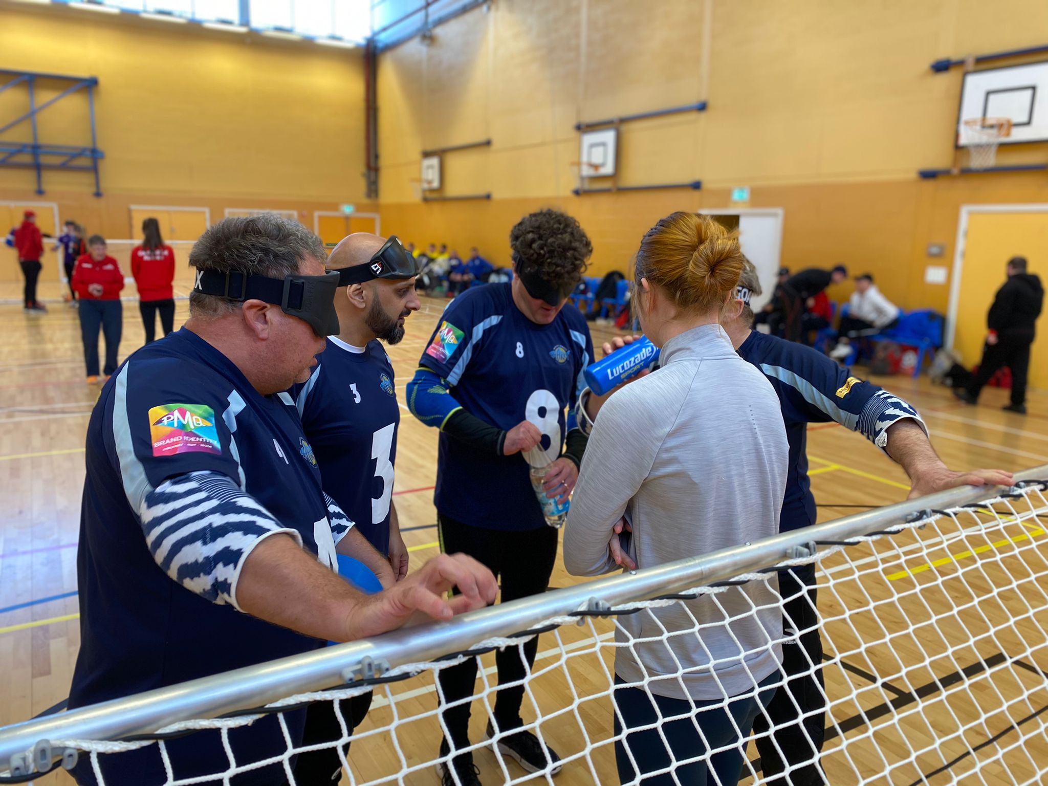 Connie, coach for Mersey Sharks is on court speaking to her team at a timeout, with players in their navy blue jerseys listening in and drinking fluids.