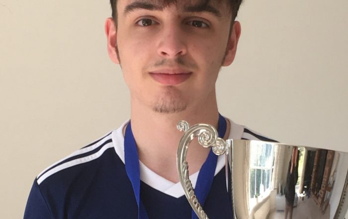 Marcus Bu-Rashid standing in front of a wall with his Winchester shirt on, holding the Intermediate Trophy and wearing his medal.