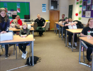 South Wales members sat in a classroom whilst taking part in a 1st aid course. The 1st aid instructor is sat in the middle of two rows of tables.