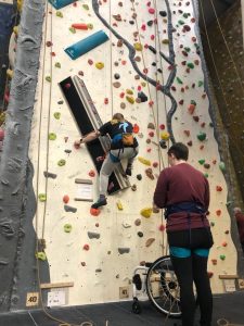 Connor facing a rock climbing wall relaying rope as someone is climbing up.